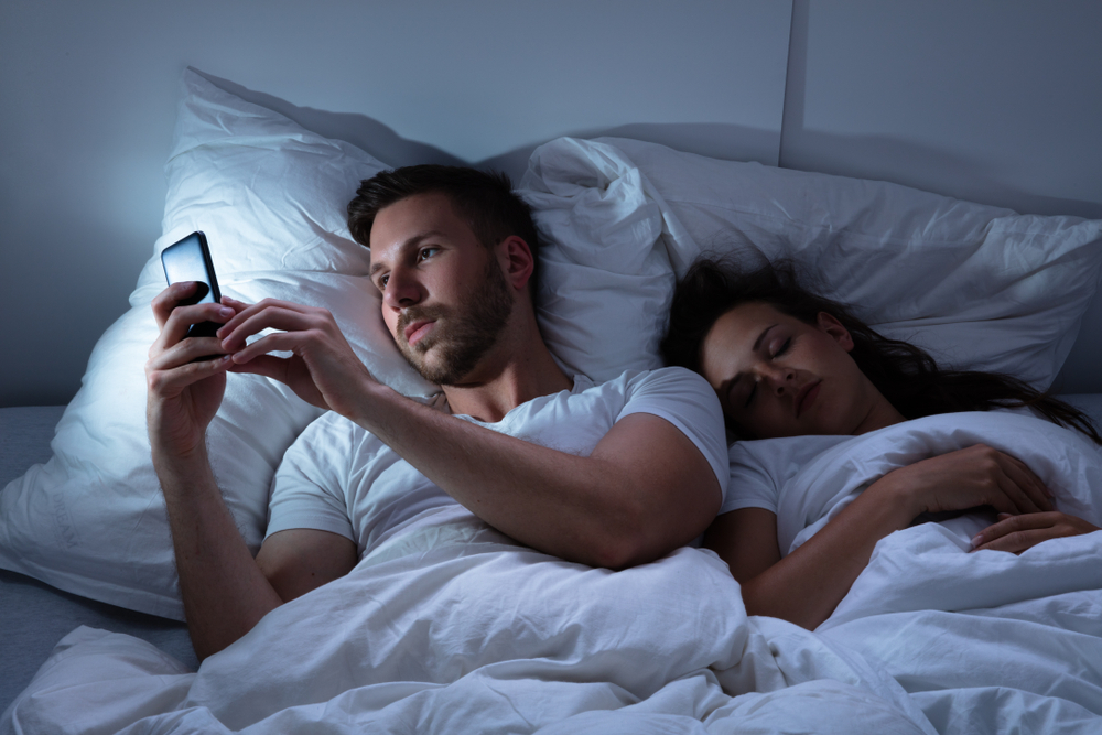 man in bed on his smartphone next to his sleeping wife