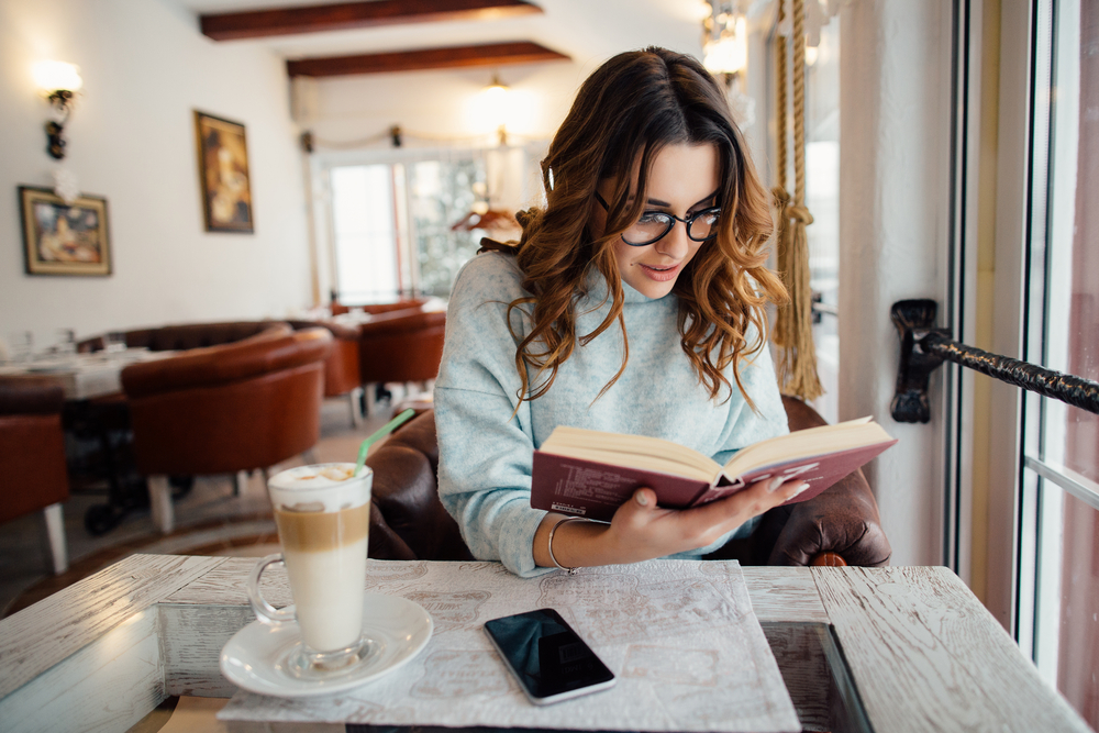 young woman reading in a cafe to complete list of Books to Read Before You’re 30