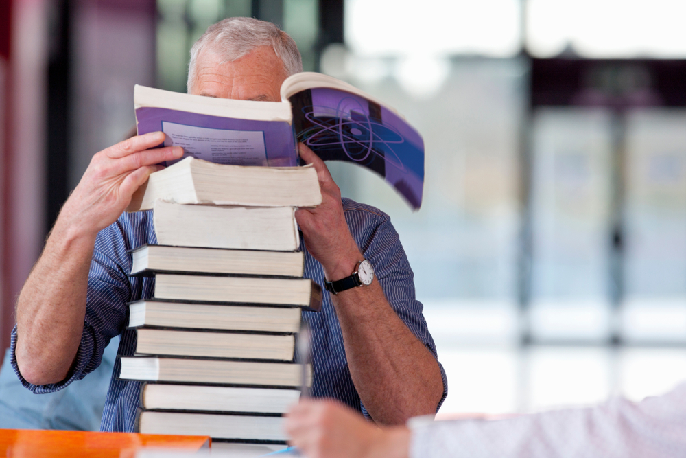 photo of a man with his face covered by an open book atop a big stack of books while he considers continuing education