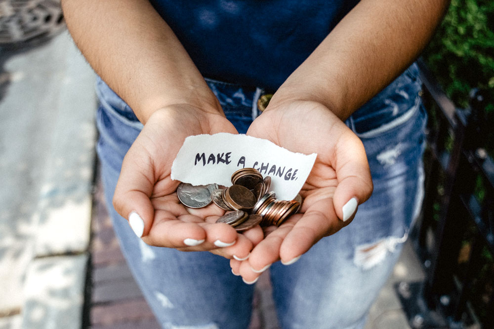 a close-up photo of a person's two hands holding up some change with a piece of paper that says "MAKE A CHANGE"