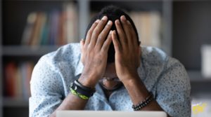 A man sitting at a desk with his hands on his head, appearing stressed or overwhelmed, with bookshelves blurred in the background.