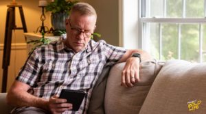A man sitting on a couch by a window, reading from a tablet, with a thoughtful expression in a cozy and well-lit living room.