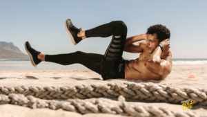 A fit man performing bicycle crunches on a sandy beach, with battle ropes in the foreground and scenic mountains and ocean in the background.