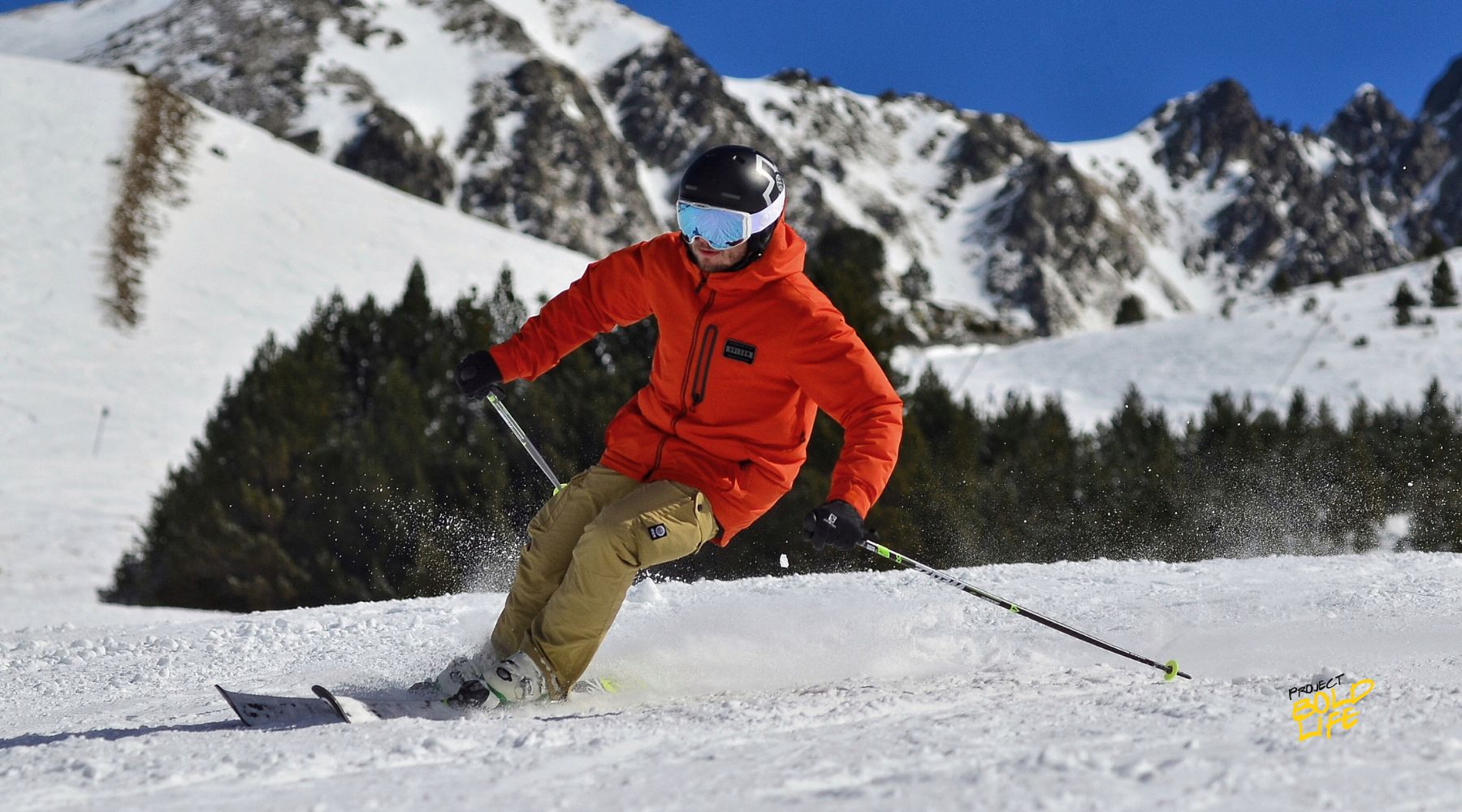 A skier in a bright orange jacket carving through fresh snow on a mountain slope, surrounded by majestic snowy peaks and evergreen trees.