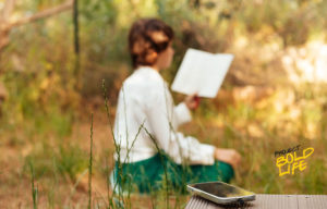 A woman reading a book outdoors with a smartphone in the foreground
