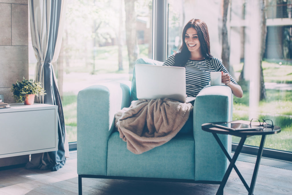 woman working from home, sitting in a chair with a blanket and computer on her lap and coffee in hand