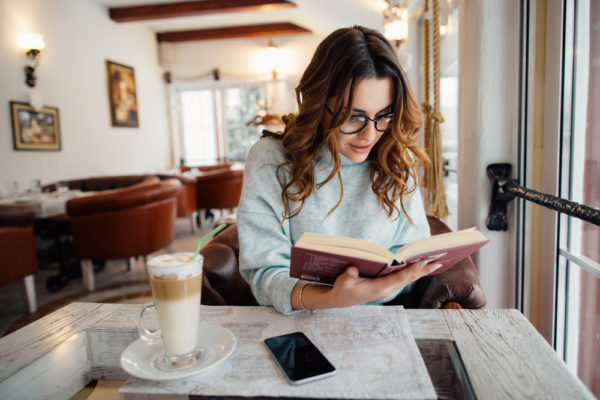 young woman reading in a cafe to complete list of Books to Read Before You’re 30