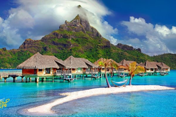 A serene tropical island retreat featuring overwater bungalows with thatched roofs, a small white sand island with palm trees and a hammock, surrounded by turquoise waters and lush green mountains under a partly cloudy sky.