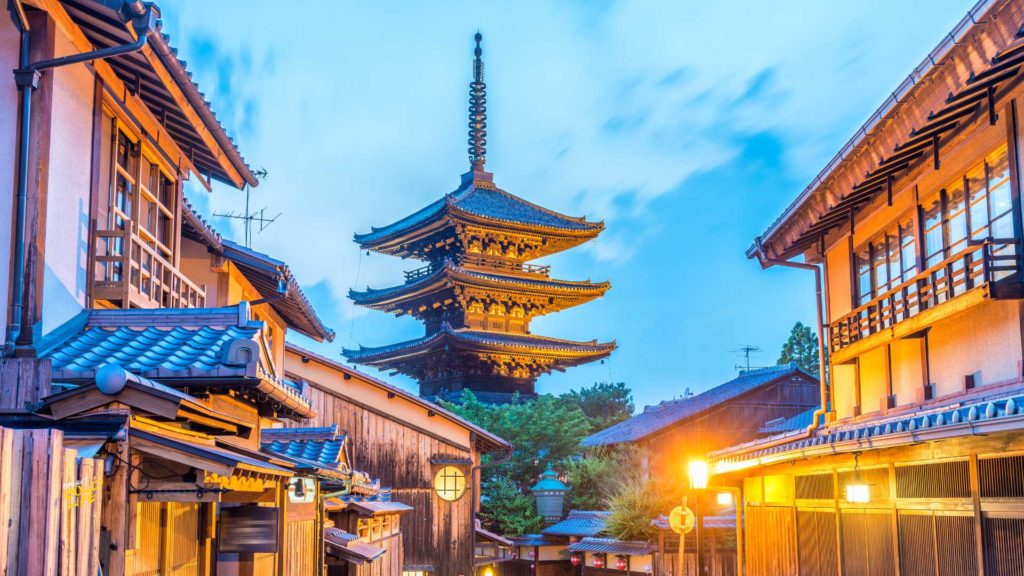 The Yasaka Pagoda, also known as Hōkanji Temple, in Kyoto, Japan, silhouetted against a colorful twilight sky, framed by blurred cherry blossoms and traditional wooden buildings illuminated by warm lanterns.